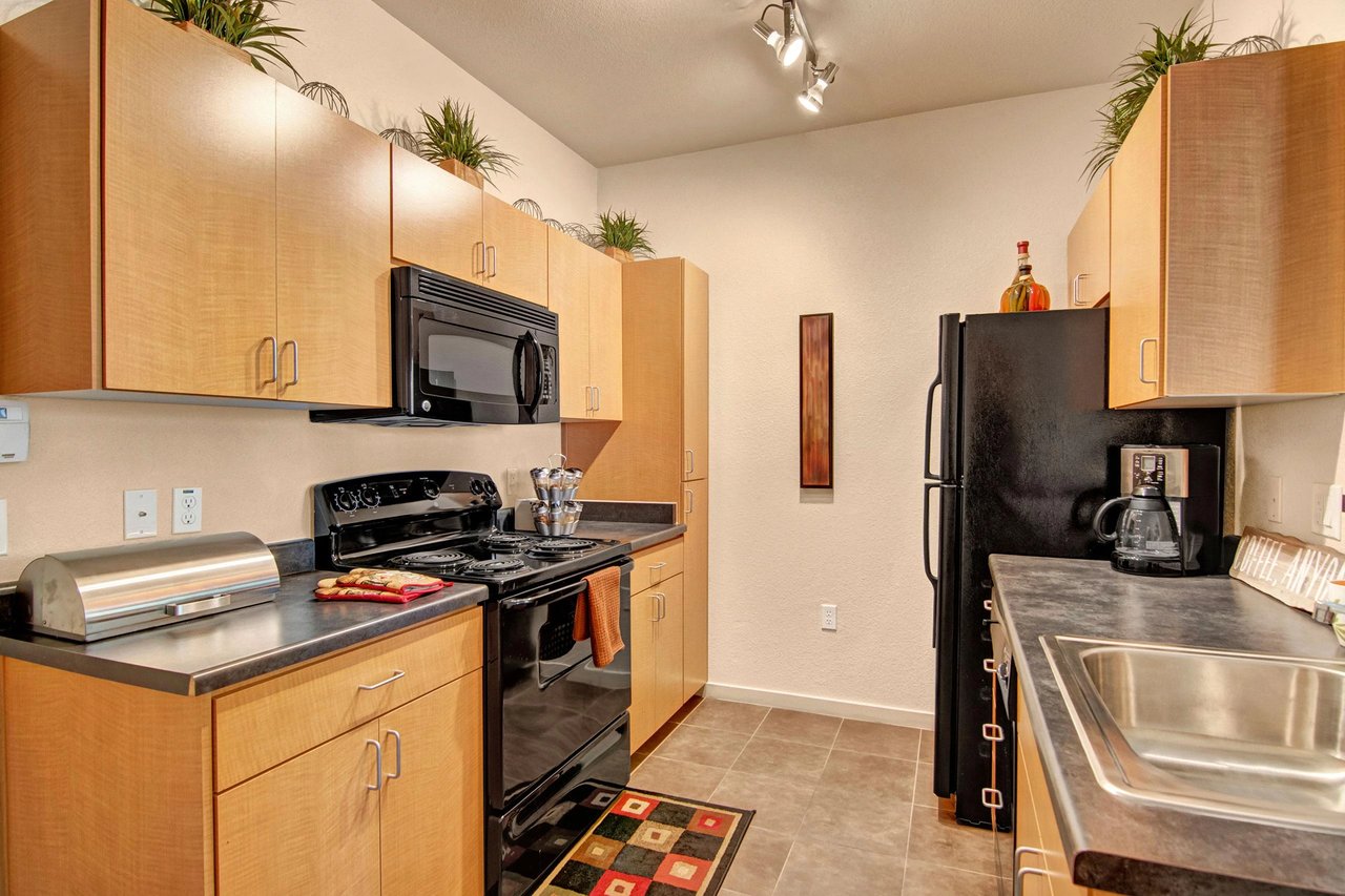 Modern kitchen with light wood cabinets, black appliances including a microwave and stove, and a stainless steel refrigerator.
