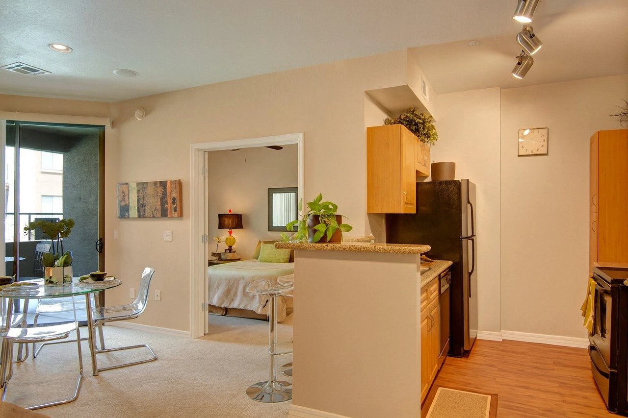 Studio apartment living area with kitchen island, dining table, and bedroom visible through doorway.