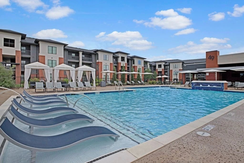 Resort-style pool with lounge chairs and cabanas in front of an apartment building.