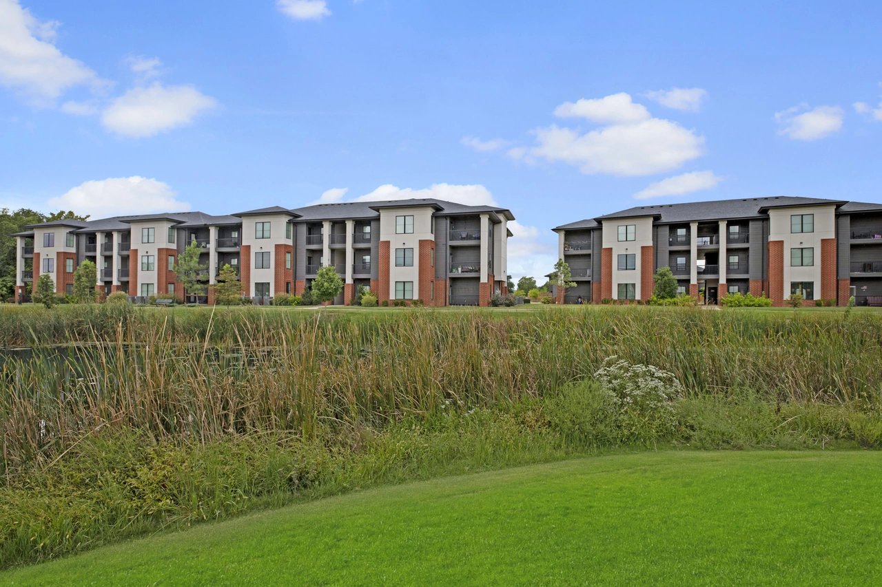 Exterior view of apartment buildings with balconies overlooking a pond and grassy area.