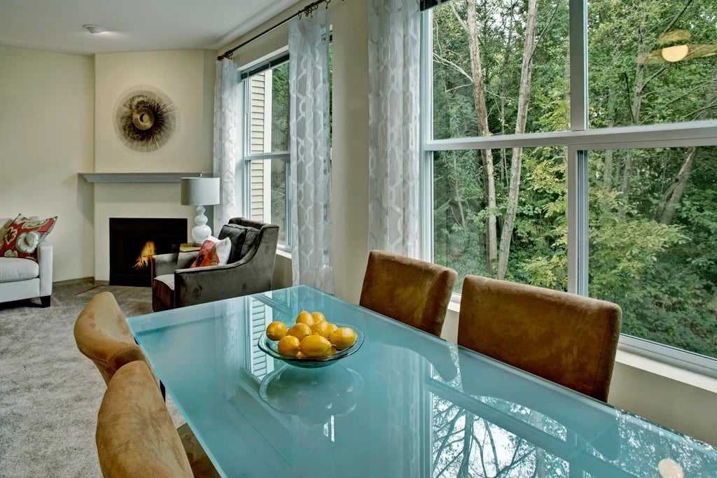 Dining area with a glass table and chairs, fireplace, and large windows overlooking trees.