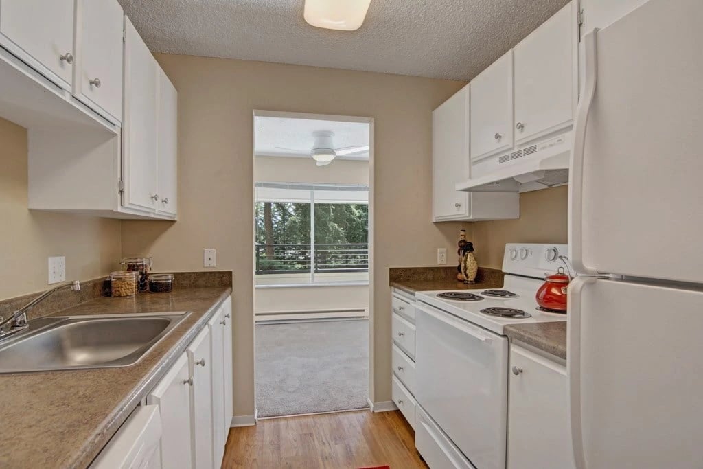 Galley kitchen with white cabinets, stainless steel sink, and white appliances. Opens to a living area.