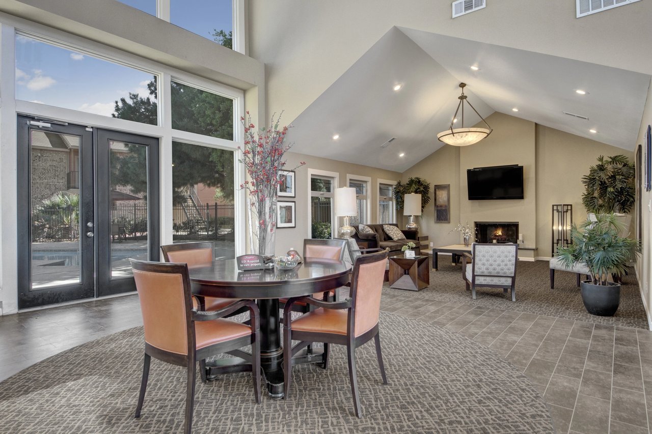 Community lounge area with a round table, chairs, fireplace, TV, and large windows overlooking the pool.