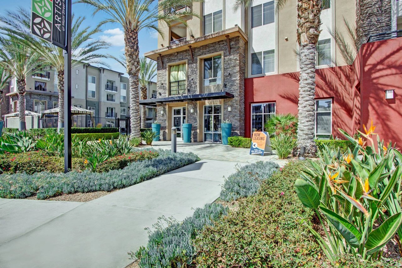 Exterior entrance of a multifamily property with palm trees, lush landscaping, and a 