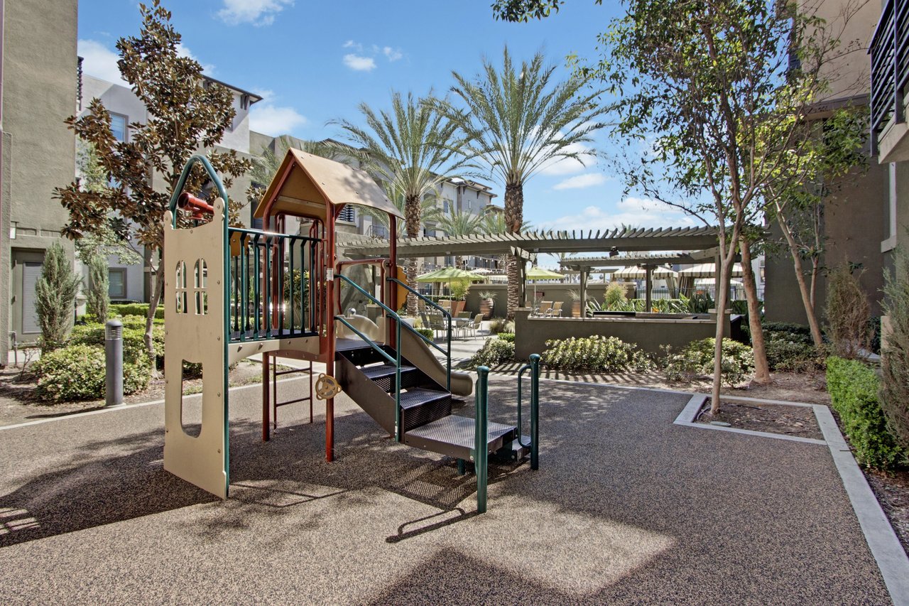 Playground area with slide and climbing structure, surrounded by trees and buildings.