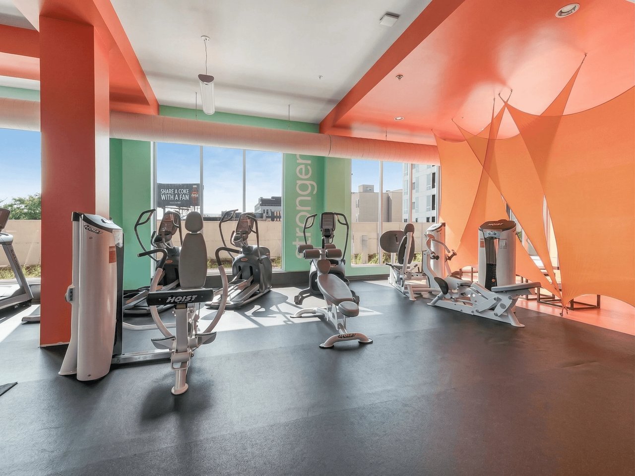 Gym with cardio machines and weight equipment, large windows, and colorful decor.