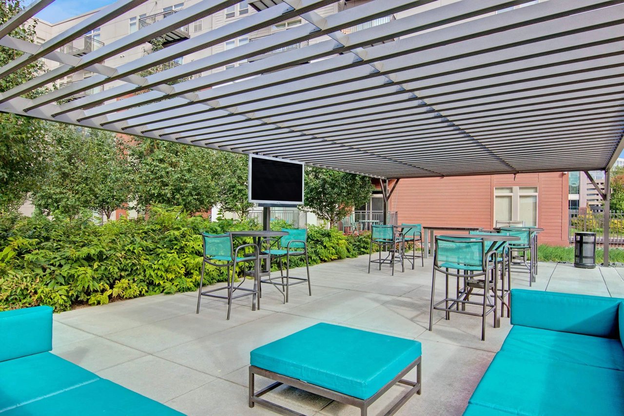 Outdoor community lounge with turquoise seating, high-top tables, planters, and a wall-mounted TV under a metal pergola.
