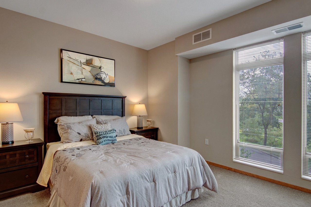 Bedroom interior with a dark wood headboard, two nightstands, lamps, and large window with blinds.