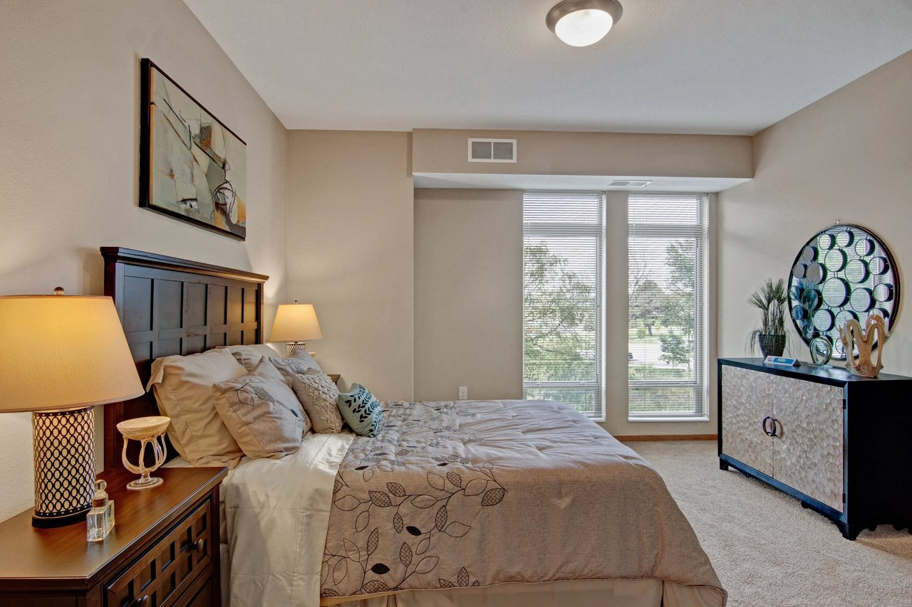 Bedroom in an apartment featuring a large bed, two nightstands, and a dresser by two large windows.