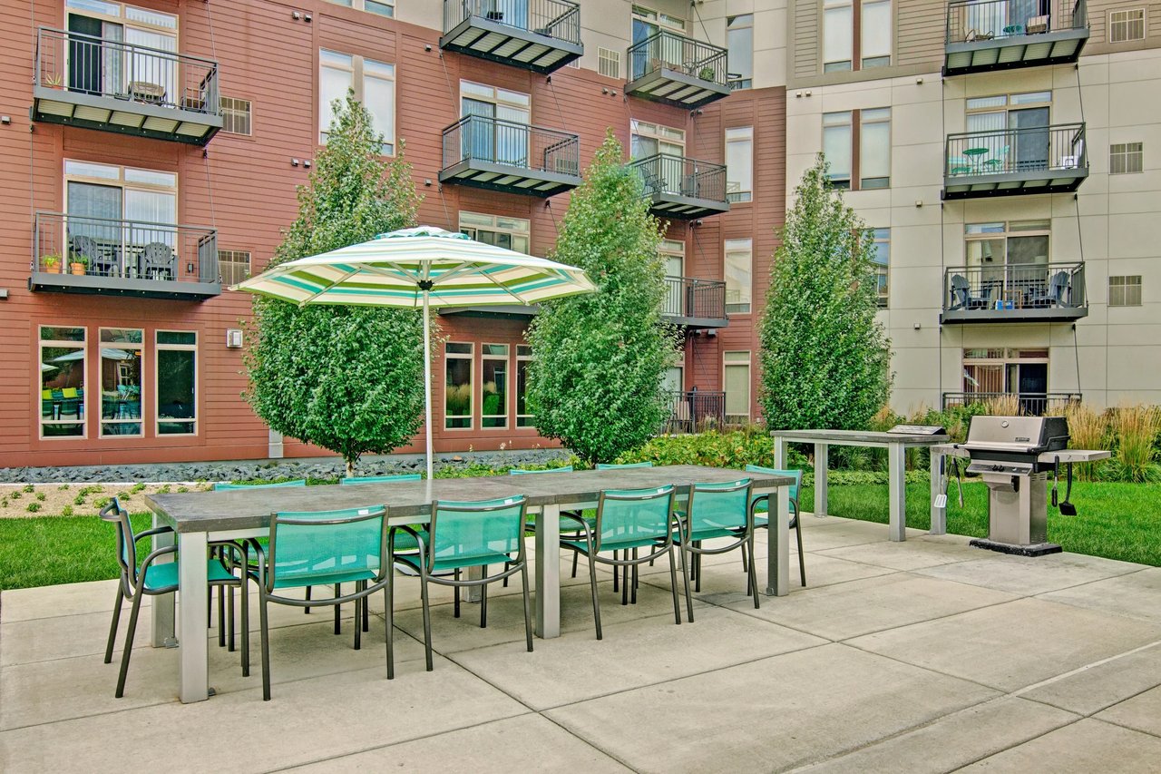 Outdoor communal patio with a long table, turquoise chairs, umbrella, grill, and apartment balconies.