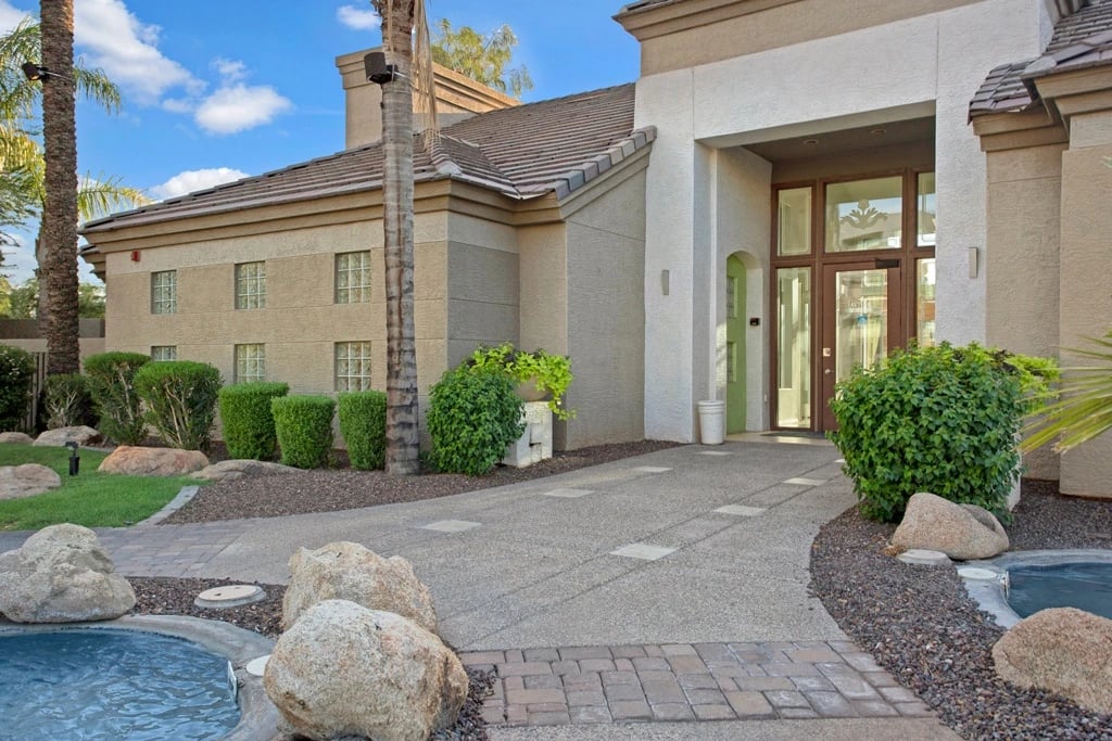 Exterior of apartment building entrance with a walkway, landscaping, and a pool.