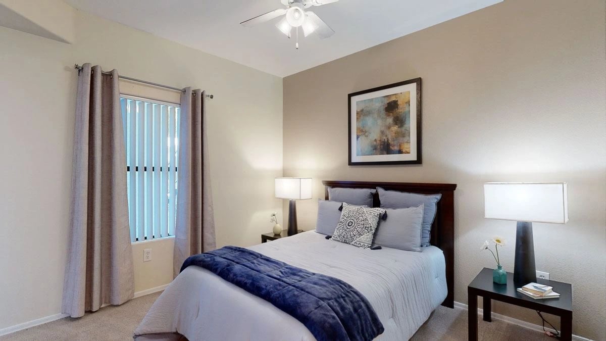 Cozy bedroom with a queen-sized bed, blue and white bedding, nightstands with lamps, and a window with blinds.