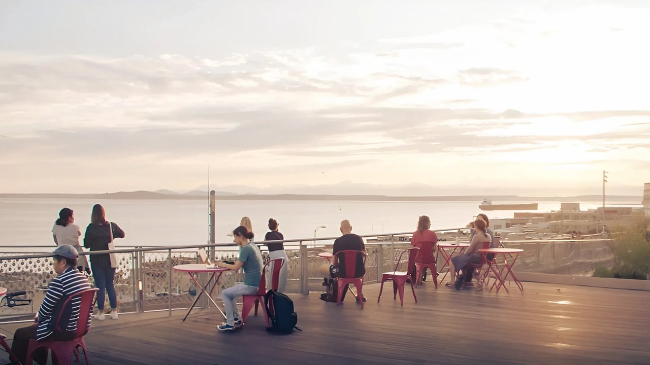 People relaxing at tables on a rooftop patio with a view of the water and sky.