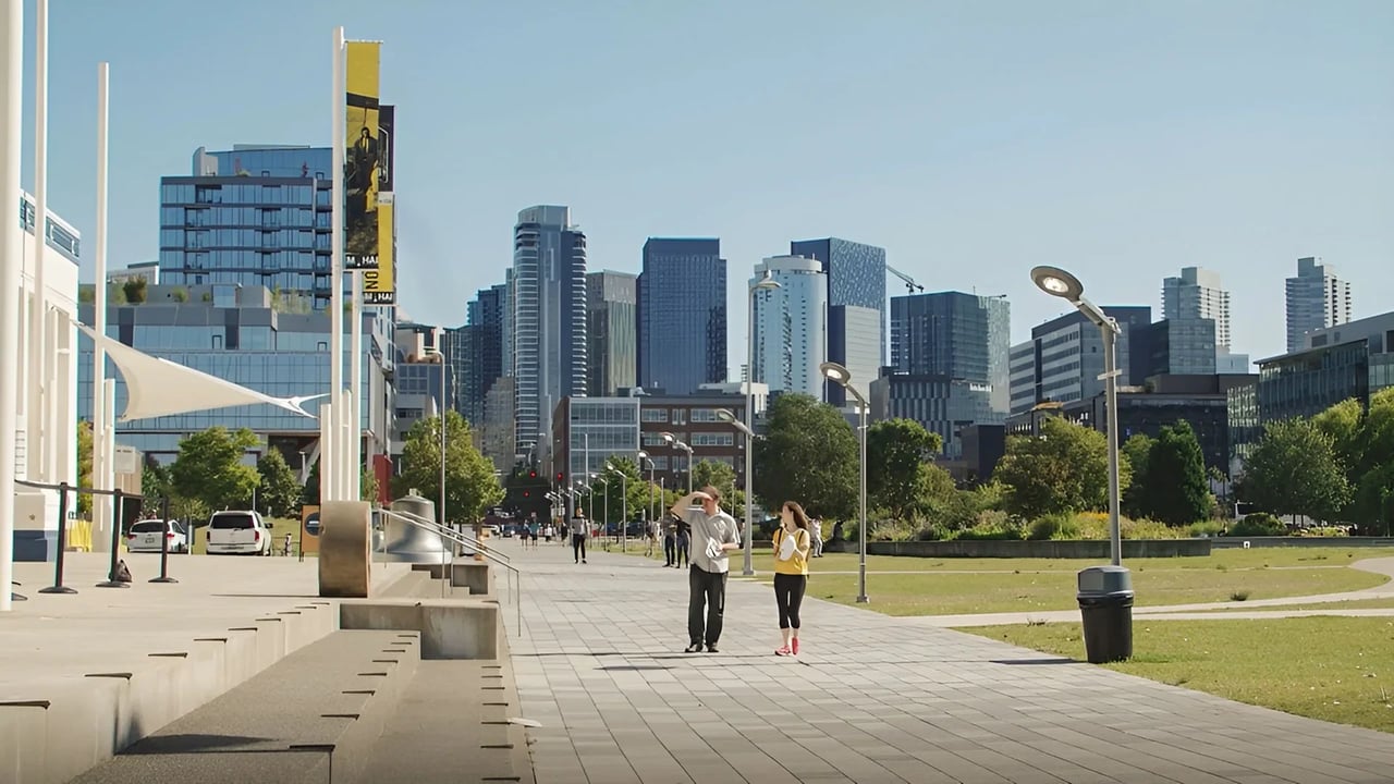 A couple walks along a paved path with a cityscape in the background.