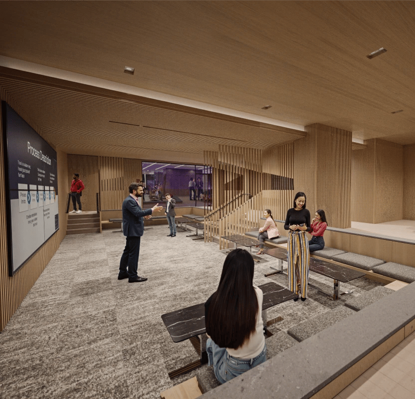 A presenter addresses a group of people seated in a modern, tiered lounge area with a large screen displaying information.