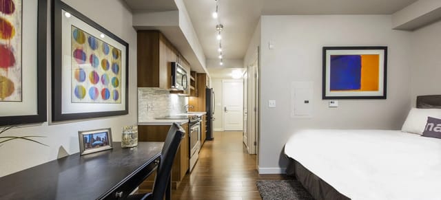 A modern studio apartment featuring a bed, desk, kitchen, and hallway.