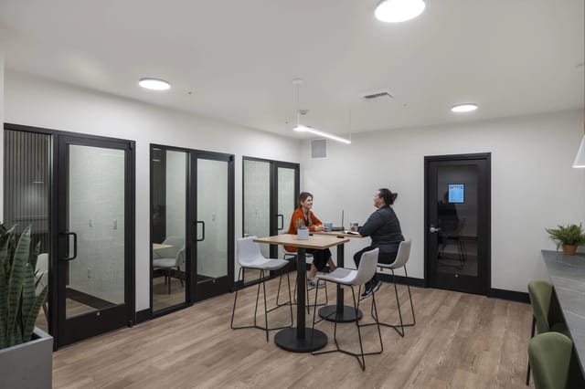 Two women collaborate at a high-top table in a shared office space with glass-walled meeting rooms.