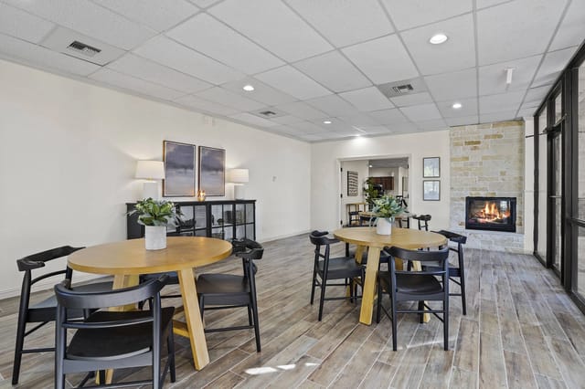 Interior community lounge with round wooden tables, black chairs, and a stone fireplace.