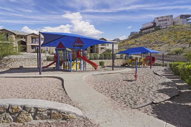 Outdoor playground with colorful equipment under blue shade canopies in a residential apartment community courtyard.