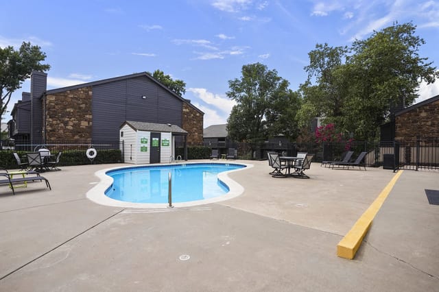 Outdoor apartment community pool with lounge chairs, tables, and a surrounding fence.