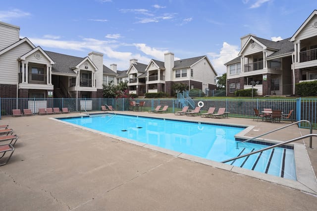 Outdoor apartment community pool with blue fence, lounge chairs, and beige multi-unit buildings in the background.