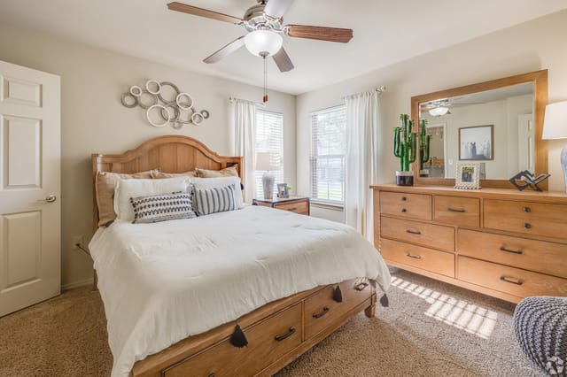 Bedroom with wooden bed, matching dresser, large mirror, ceiling fan, and two windows with white curtains.