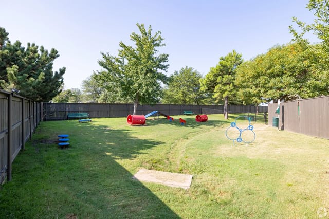 Fenced grassy community playground with red tunnels, a slide, and other play equipment.