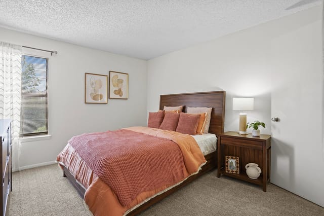 Cozy bedroom with a queen-size bed and plush bedding, featuring a nightstand with a lamp and a window with blinds.