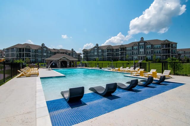 Outdoor community pool with blue-tiled deck, lounge chairs, and apartment buildings in the background.