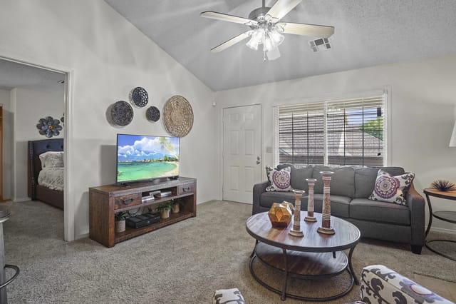 Living room in an apartment featuring a gray sofa, round coffee table, TV stand, ceiling fan, and window.