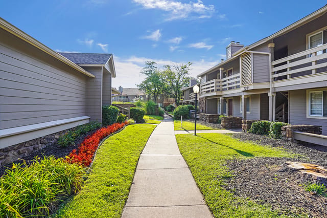 A paved walkway leads through a grassy courtyard with manicured flower beds between apartment buildings.