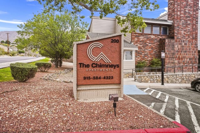 Exterior sign for The Chimneys Apartments with brick building and landscaping.