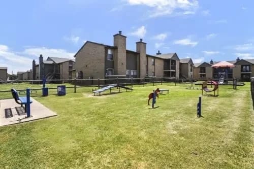 Outdoor dog park with agility equipment and apartment buildings in the background.