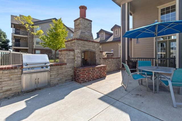 Outdoor grilling station with a built-in grill, stone fireplace, and seating area under a blue umbrella.