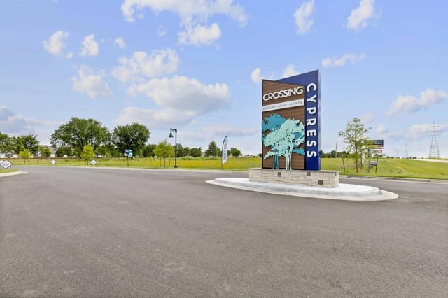 Entrance sign for Crossing Cypress Luxury Apartments at a roundabout, with landscaping and blue sky.