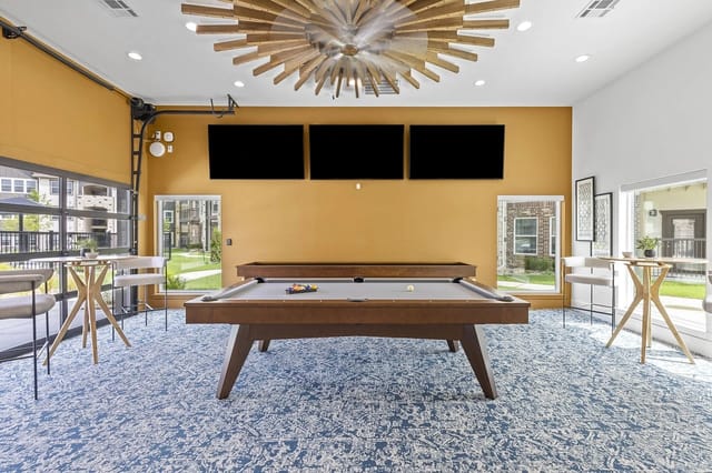 Interior of a community lounge with a pool table, three wall-mounted TVs, and large windows.