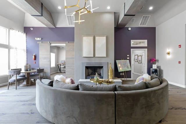 Modern communal living area with curved gray sofa, fireplace, purple accent wall, and dining table.