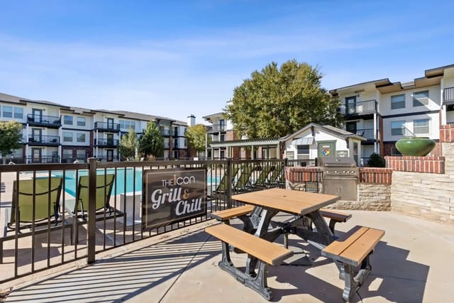 Outdoor community grill area with a picnic table and pool in the background.