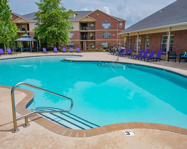 Outdoor pool at apartment community with blue water, purple lounge chairs, and brick buildings.