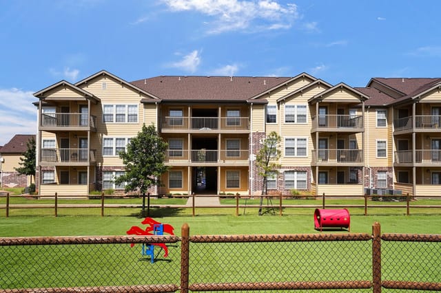 Exterior view of beige apartment buildings with balconies and a fenced grassy courtyard.