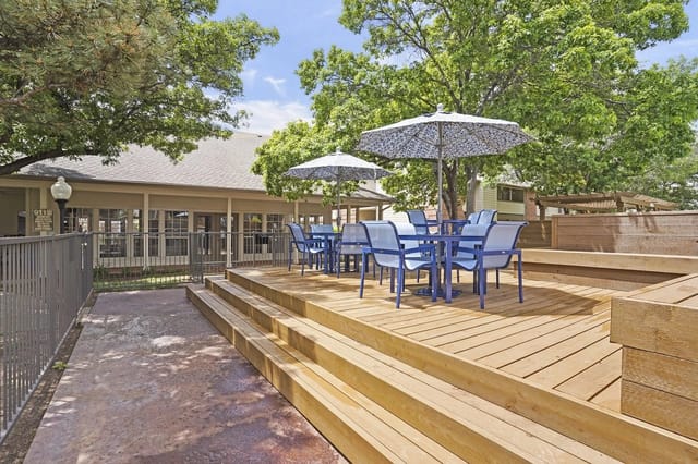 Outdoor community deck with blue chairs, umbrellas, steps, and railing under trees.