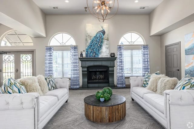 Community lounge with white tufted sofas, a dark fireplace, peacock artwork, and arched windows.