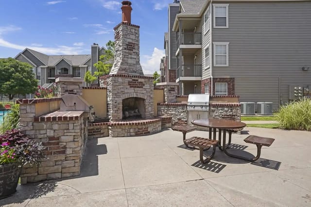 Outdoor community grilling area with stone fireplace and table, surrounded by apartment buildings.