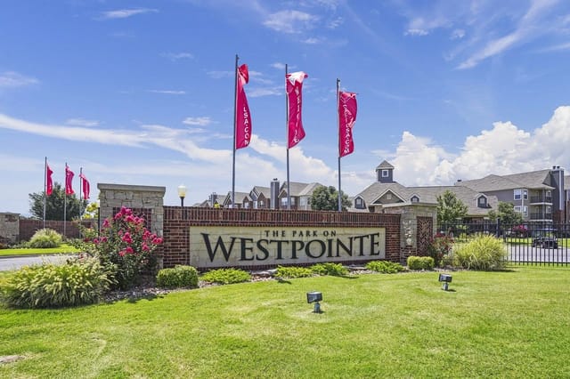 Entrance sign for The Park on West Pointe with brick pillars, landscaping, and red flag banners.