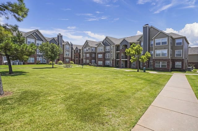 Exterior view of a multi-building apartment community with a green lawn, trees, and a sidewalk.
