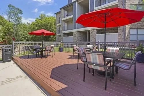Outdoor community patio with tables, chairs, and red umbrellas on a wooden deck beside an apartment building.