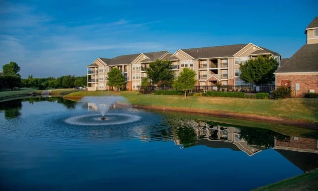 Exterior view of a multi-family apartment community with a pond, fountain, and landscaped grounds.