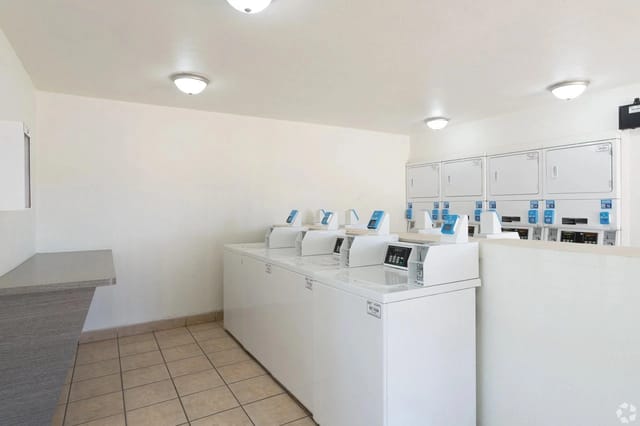 Communal laundry room with row of white top-load washers and matching dryers.
