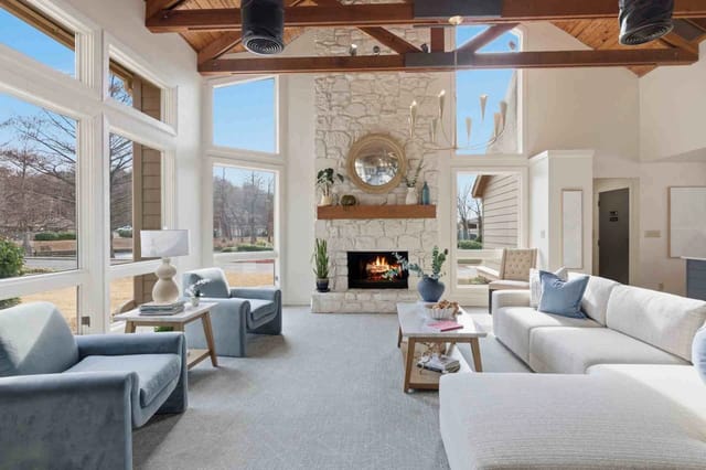 Bright living room with a stone fireplace, vaulted wooden beams, and large windows.