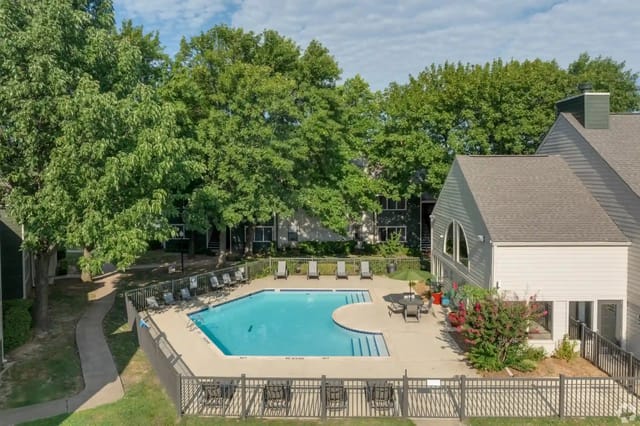 Outdoor pool area with lounge chairs around a blue pool, shaded by large trees.